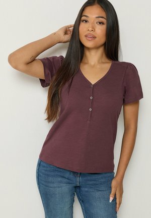 Mujer con cabello largo y oscuro que lleva una camiseta morada de manga corta con botones y vaqueros azules, posando con una mano detrás de la cabeza.