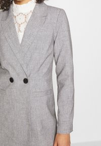 Szara dwurzędowa marynarka z teksturowanej tkaniny, z czarnymi guzikami, klapami oraz kieszeniami po bokach. Pod spodem widoczna koronkowa bluzka.