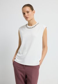 Eine Frau mit zurückgegeltem Haar trägt ein weißes, ärmelloses Oberteil mit verziertem Ausschnitt und mauvefarbenen Hosen und steht mit den Händen in den Taschen.