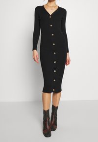 Schwarzes, geripptes Kleid mit langen Ärmeln, V-Ausschnitt und goldenen Knopfakzenten auf der Vorderseite. Enganliegende Form mit einem knielangen Saum.