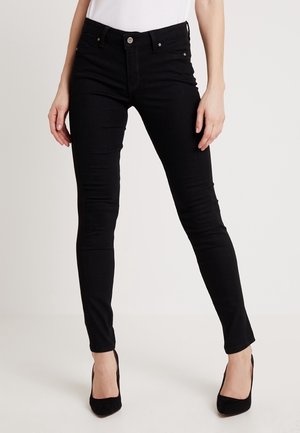 Vrouw draag slim-fit zwarte jeans en zwarte hoge hakken, staand tegen een effen lichte achtergrond.