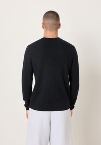 Schwarzer Strickpullover mit rundem Ausschnitt, langen Ärmeln und weicher Textur, getragen über grauem Jogginghose. Ansicht von hinten.