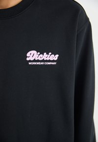 Felpa nera con logo ricamato rosa "Dickies Workwear Company" sul lato sinistro del petto. Realizzata in un tessuto strutturato con scollatura a girocollo.
