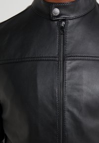 Jachetă din piele neagră cu guler înalt, fermoar frontal și detalii cusute. Are o textură netedă și accente hardware minime.