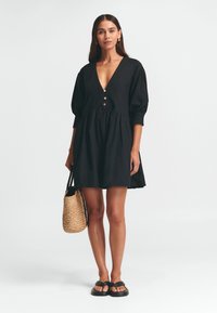 Schwarzes Kleid mit tiefem V-Ausschnitt und Knopfdetails, ausgestattet mit Puffärmeln und einem ausgestellten Rock. Begleitet von einer geflochtenen Tasche und Flip-Flops.