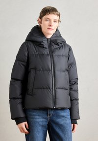 Doudoune noire avec capuche, présentant des sections matelassées horizontales, une fermeture éclair à l'avant et des poignets en tricot côtelé. Portée par-dessus un jean bleu.
