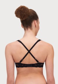 Black lace bra with crisscross straps, featuring gold hardware accents and a delicate floral pattern along the band. Back view displayed.