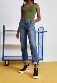 Top verde, jeans di denim blu a vita alta con risvolti, e sneakers nere alte con accenti arancioni. Sullo sfondo c'è un carrello metallico blu.
