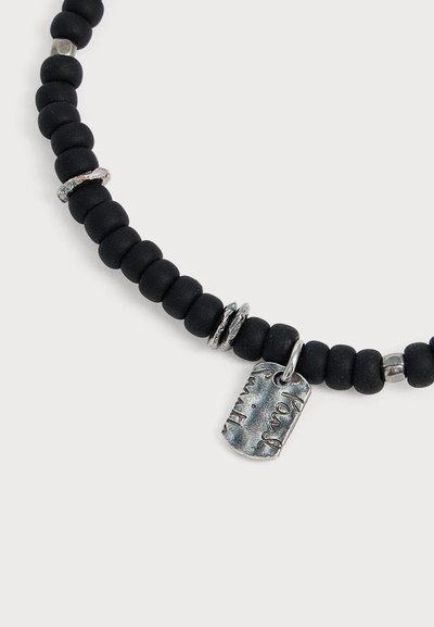 Collier en perles noires avec petits anneaux en argent et pendentif rectangulaire en argent gravé d’une écriture cursive.
