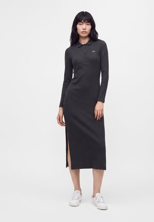 Donna che indossa un lungo vestito nero con colletto, spacco laterale e sneakers bianche, in piedi davanti a uno sfondo bianco.
