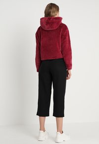 Hoodie corto in pelliccia sintetica bordeaux con cappuccio, abbinato a pantaloni cropped neri e sneakers bianche chunky, mostrato da dietro.