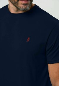 Tumši zila īsām piedurknēm T-kreklā no gludas auduma, kurā ir neliels sarkans izšūts dizains kreisajā krūšu pusē.