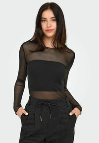 Młoda kobieta ubrana w czarną, przejrzystą bluzkę z długim rękawem na czarnym topie typu tube oraz spodniach z paskami w jodełkę, z rękami w kieszeniach.