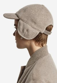 Ljusbrun ullkeps med rundad krona, styv skärm och öronlappar. Har en broderad logotyp på kepsen och ribbat tyg vid kragen.