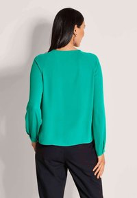 Grüne Seidenbluse mit langen Ärmeln, gerade geschnitten und mit knöpfbaren Manschetten. Glatte Textur und minimalistisches Design ohne Muster.