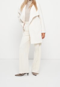 Lång vit kappa med en draperad krage, beige polokrage och vita vida byxor. Bruna spetsiga klackar och neutrala färger. Slät textur.