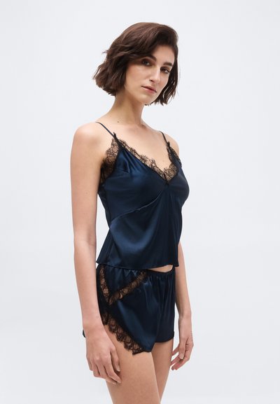 Žena oblečená v námornícky modrom saténovom tielkovom topi a zodpovedajúcich šortkách s čiernym krajkovým lemom, stojaca pred jednoduchým svetlosivým pozadím.