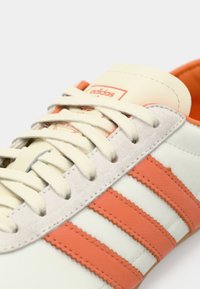 Λευκό αθλητικό παπούτσι με τρεις πορτοκαλί ρίγες, λεπτομέρειες από ανοιχτό γκρι σουέντ και κρέμ κορδόνια. Το εσωτερικό είναι πορτοκαλί με ετικέτα λογότυπου Adidas.
