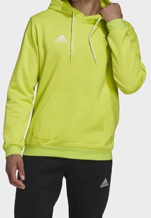 Personne portant un sweat à capuche Adidas jaune vif avec des cordons blancs et un pantalon Adidas noir, ajustant le col du sweat à capuche d'une main.