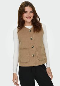 Giovane donna con lunghi capelli castani che indossa un gilet beige abbottonato sopra una camicia bianca a maniche lunghe e pantaloni neri, sorridente e in posa.