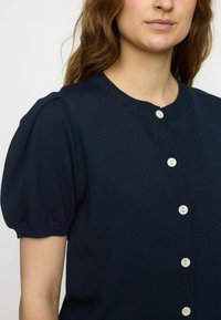 Marineblå strikket cardigan med korte pufærmer, rund halsudskæring og hvide knapper, der løber ned foran. Glat tekstur.