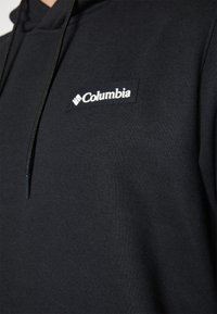 Juodas gobtuvu džemperis, pagamintas iš medvilnės mišinio, su baltu Columbia logotipo pavadinimu krūtinėje ir juodais raišteliais.