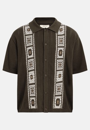 Tumši olīvu krāsas īsroku trikotāžas krekls ar pogām priekšpusē, apkakli un baltiem ģeometriskiem kvadrātu rakstiem, kas iet pa vertikāli abās pusēs.