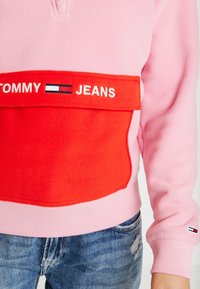 Różowy sweter polarowy z dużą czerwoną kieszenią, w której wyszyto "TOMMY JEANS" na biało. Rękawy i dół wykończone ściągaczem. Noszony z niebieskimi, przetartymi dżinsami.