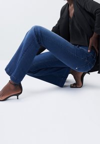 Femme vêtue d'un blazer noir, d'un haut foncé et d'un jean bleu accroupie, portant des sandales noires à talons hauts sur un fond blanc.