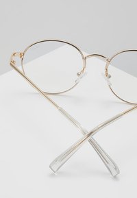Lunettes à monture en métal doré avec des verres ronds, des plaquettes de nez transparentes et des branches transparentes qui se croisent à la base. Design minimaliste et léger.