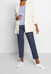 Blazer blanc croisé porté sur un top bleu clair, associé à un pantalon en denim foncé retroussé et des baskets blanches. Tissu lisse, coupe ajustée.