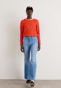 Rød strikket genser, lyseblå vide jeans, svarte sneakers. Modellen har på seg en mørk caps med et flerfarget logo og hvite sokker.