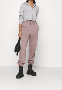 Sudadera gris recortada con cremallera, pantalones de chándal rosas con cordón y botas de combate negras con suelas gruesas, sosteniendo una pequeña bolsa negra.
