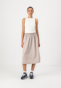 Haut sans manches blanc avec logo rose, associé à une jupe beige longueur cheville avec une taille élastique. Le modèle porte des baskets bleues.