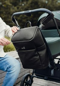 Černý kožený batoh s prošitou texturou, přihrádkami na zip a nastavitelné popruhy. Připevněný k rukojeti kočárku, ukazující praktičnost.