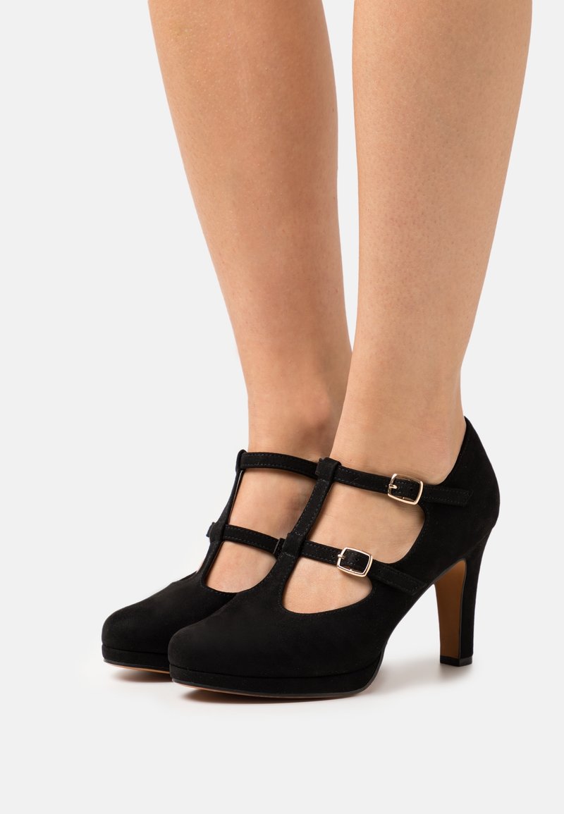 Escarpins à talons hauts en daim noir, à bride en T, avec des fermetures à double boucle, portés sur une paire de pieds sur un fond clair uni.