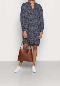 Robe bleu marine avec un motif paisley, décolleté en V et manches longues. Un sac fourre-tout en cuir marron et des baskets blanches complètent la tenue.