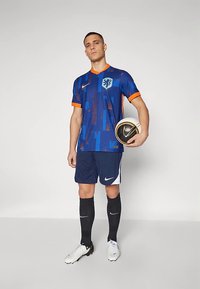 Mees sinises ja oranžis jalgpallivormis, hoides musta-valget Nike jalgpalli, seistes ühtlase tausta ees.