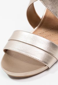 Gros plan d'une sandale beige métallisée montrant la bride d'orteil, la semelle intérieure rembourrée avec des perforations et la texture lisse.