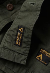 Olijfgroene utility shirt stof, met een bruine knop, twee zwarte labels met gele tekst en een gestructureerde afwerking. Accenten omvatten stikseldetails.