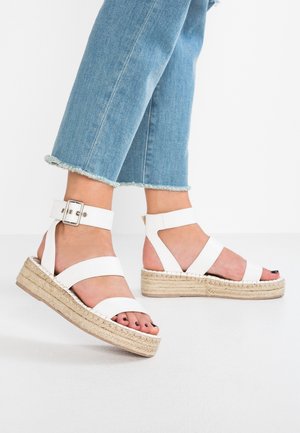 White espadrille sandals with wide straps and ankle buckle, featuring a woven jute sole, set against light blue denim cropped jeans.
