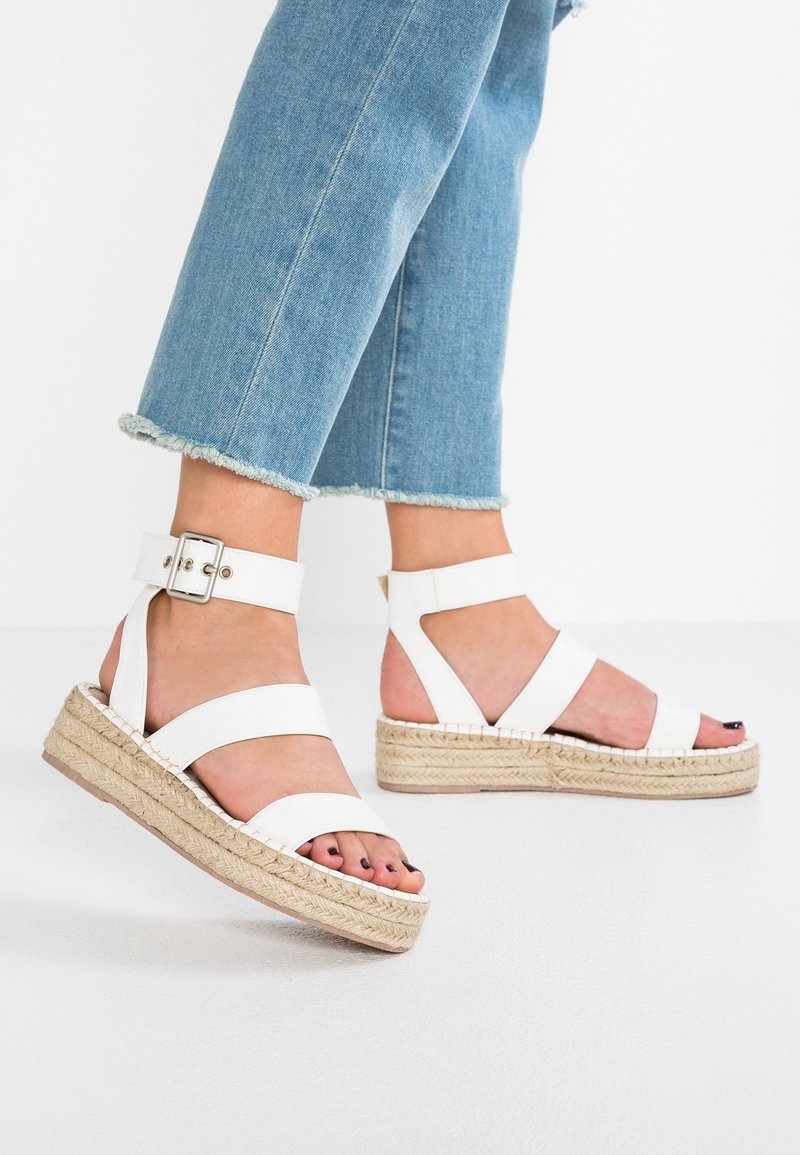 Sandales espadrilles blanches à larges sangles et boucle de cheville, avec une semelle en jute tressé, posées sur un jean crop en denim bleu clair.