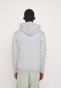 Persona de espaldas vistiendo una sudadera con capucha gris claro y pantalones cargo verde claro contra un fondo blanco liso.