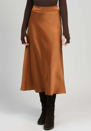 Femme portant un haut marron à manches longues, une jupe midi en satin couleur caramel et des bottes noires montantes devant un fond gris uni.