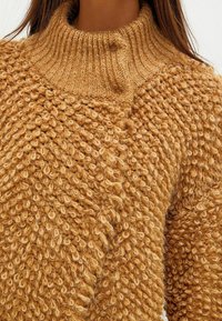 Texturiertes, senf-farbendes Sweater mit hohem Rippkragen, locker sitzend und weichem, flauschigem Strickmuster. Zwei Knopfakzente in der Nähe des Kragens.