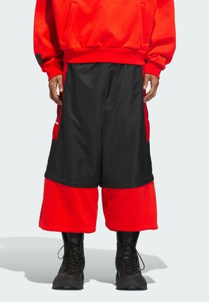 Mand iført sorte og røde brede lagdelte shorts med sorte snørebåndsstøvler og en rød sweatshirt, stående på en lys baggrund.