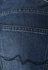 Vaqueros de denim de tono azul oscuro, con un distintivo detalle de costura ondulada y un acabado texturizado, que muestra un cosido reforzado.