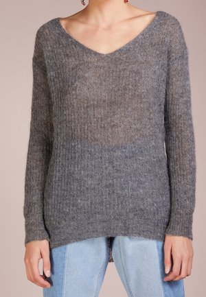 Vrouw draagt een zachte, pluizige grijze V-hals trui met lange mouwen, gecombineerd met lichtblauwe spijkerbroek tegen een effen achtergrond.