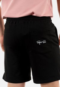 Pantalones cortos de algodón negros con una cinturilla elástica y un bolsillo trasero que presenta un texto bordado en blanco. Diseño simple, textura suave.