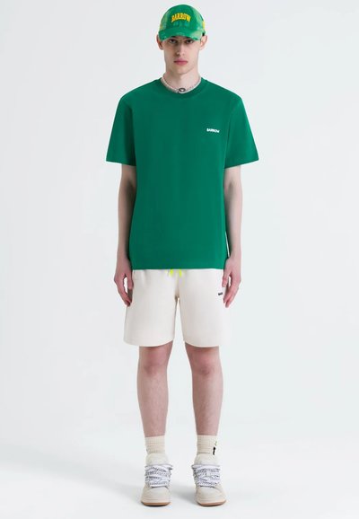T-shirt verde in cotone con logo bianco, pantaloni corti crema con coulisse gialla e sneakers beige con lacci grigi. Accentuato da un cappellino verde.
